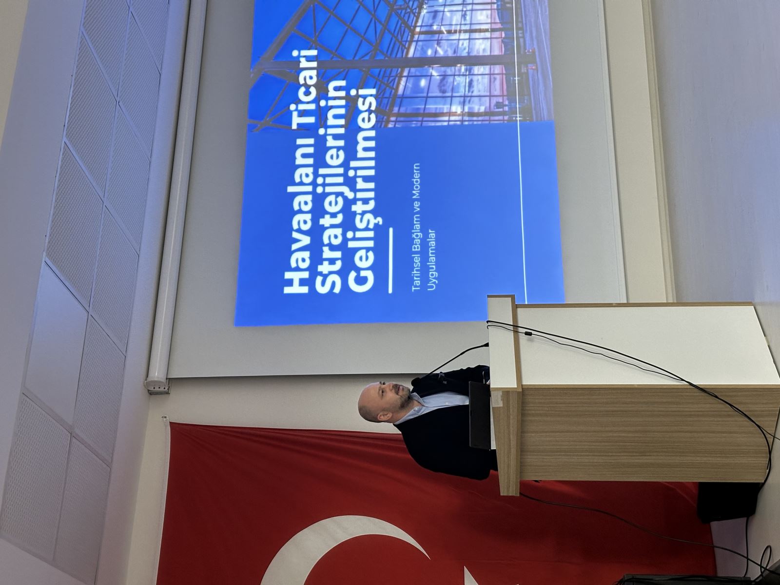 Ahmet Orçun Songur, kürsüde sunum yaparken görülüyor. Arka planda Türk bayrağı ve yansıtılan sunum slaytları var. Slaytlarda "Havacılık Dışı Gelirlerin Artırılması" ve "Havaalanı Ticari Stratejilerinin Geliştirilmesi" başlıkları yer alıyor.